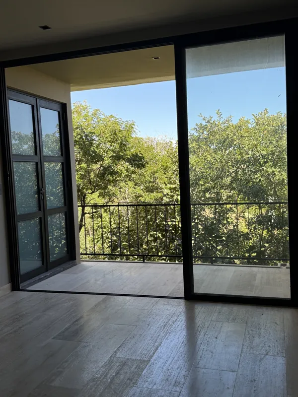 A bright, finished interior room in an Arthouse Signature one-bedroom apartment, featuring light-colored tiled floors, light grey walls, an installed air conditioning unit, and a large sliding glass door opening to a private balcony with lush gree...