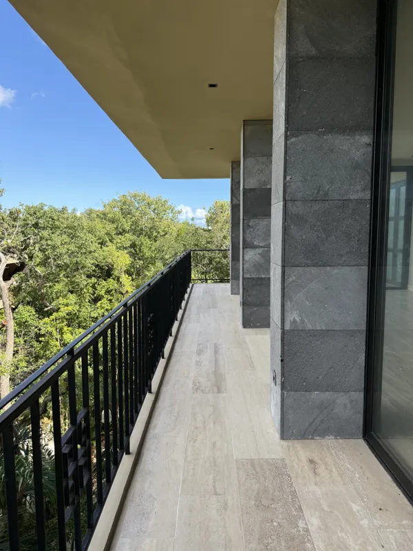 A bright, finished interior room in an Arthouse Signature one-bedroom apartment, featuring light-colored tiled floors, light grey walls, an installed air conditioning unit, and a large sliding glass door opening to a private balcony with lush gree...