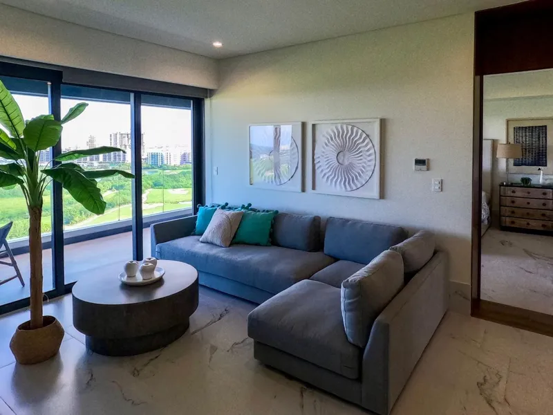 A spacious, elegantly furnished living room in a Woha residence, featuring modern decor, marble floors, and large sliding glass doors opening to a private terrace with panoramic views of the natural reserve, golf course, and distant Caribbean Sea.