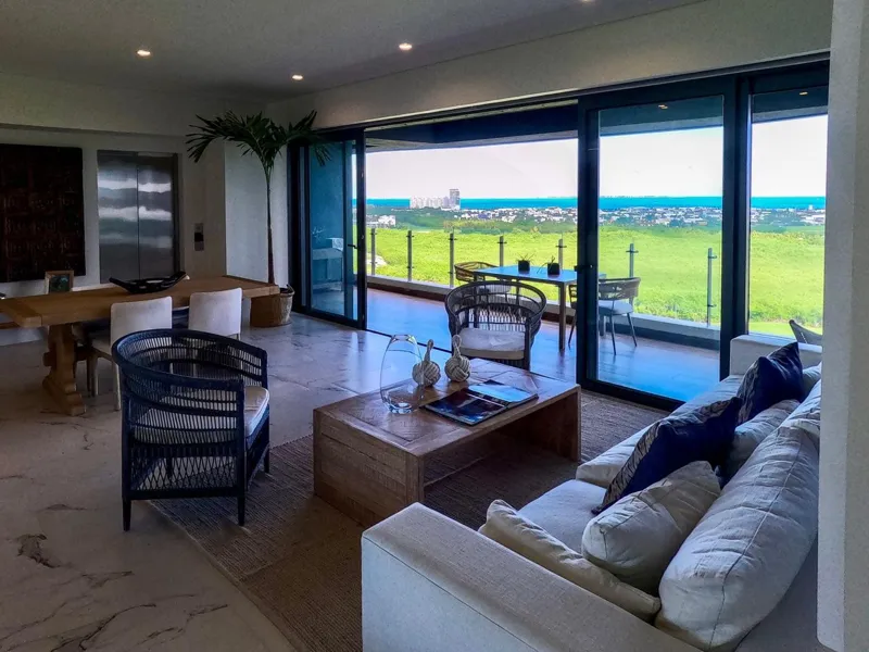 A spacious, elegantly furnished living room in a Woha residence, featuring modern decor, marble floors, and large sliding glass doors opening to a private terrace with panoramic views of the natural reserve, golf course, and distant Caribbean Sea.