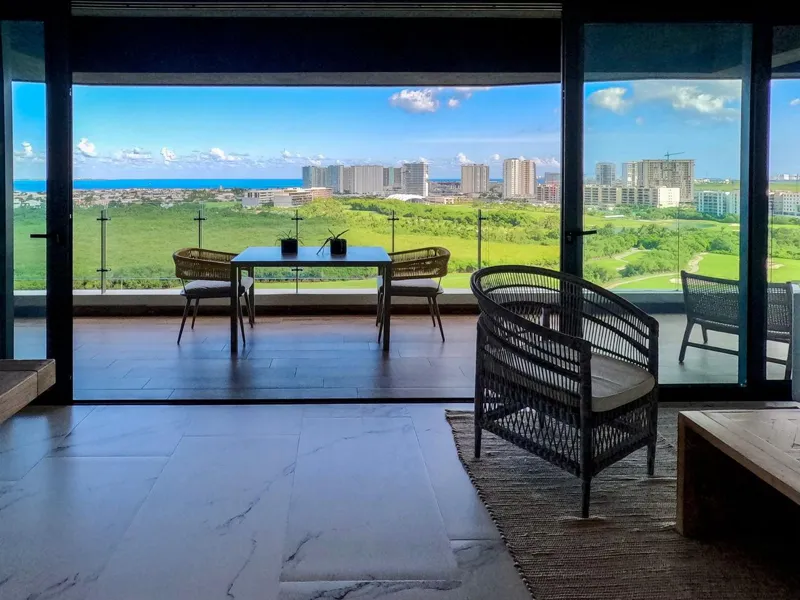 A spacious, elegantly furnished living room in a Woha residence, featuring modern decor, marble floors, and large sliding glass doors opening to a private terrace with panoramic views of the natural reserve, golf course, and distant Caribbean Sea.