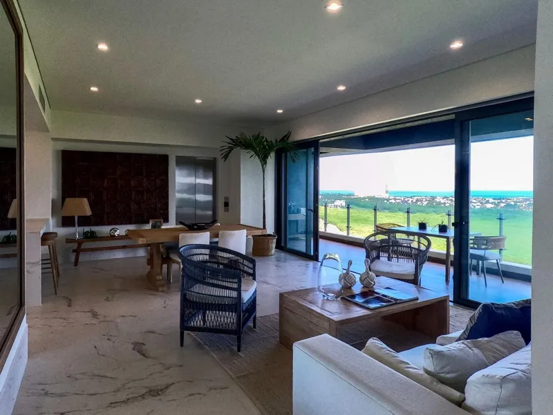 A spacious, elegantly furnished living room in a Woha residence, featuring modern decor, marble floors, and large sliding glass doors opening to a private terrace with panoramic views of the natural reserve, golf course, and distant Caribbean Sea.
