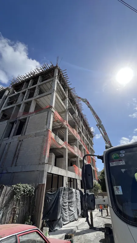 A concrete pump truck and mixer truck actively pouring concrete onto the 6th level of the Maia development building, which features a multi-story concrete structure with rebar visible at the top and orange safety netting on the lower floors, under...