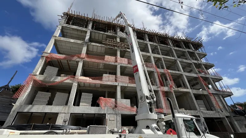 A concrete pump truck and mixer truck actively pouring concrete onto the 6th level of the Maia development building, which features a multi-story concrete structure with rebar visible at the top and orange safety netting on the lower floors, under...