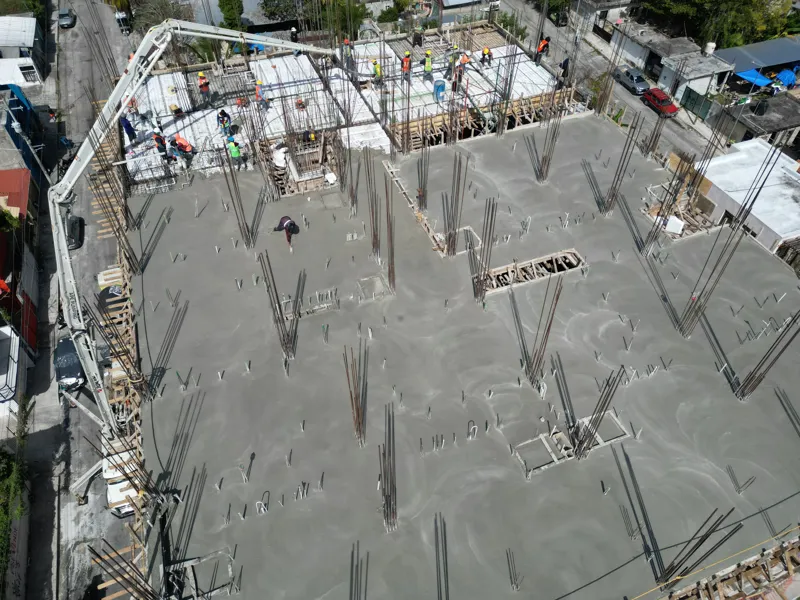 A concrete pump truck and mixer truck actively pouring concrete onto the 6th level of the Maia development building, which features a multi-story concrete structure with rebar visible at the top and orange safety netting on the lower floors, under...