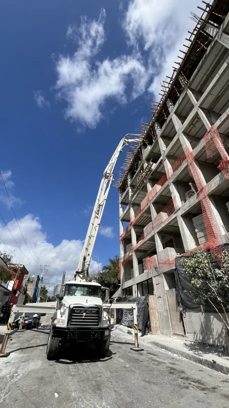 A concrete pump truck and mixer truck actively pouring concrete onto the 6th level of the Maia development building, which features a multi-story concrete structure with rebar visible at the top and orange safety netting on the lower floors, under...
