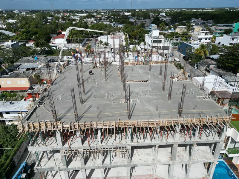 A concrete pump truck and mixer truck actively pouring concrete onto the 6th level of the Maia development building, which features a multi-story concrete structure with rebar visible at the top and orange safety netting on the lower floors, under...