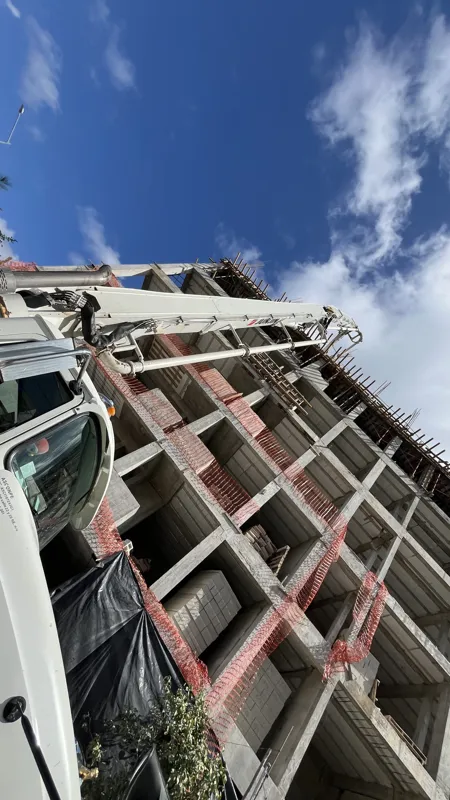 A concrete pump truck and mixer truck actively pouring concrete onto the 6th level of the Maia development building, which features a multi-story concrete structure with rebar visible at the top and orange safety netting on the lower floors, under...