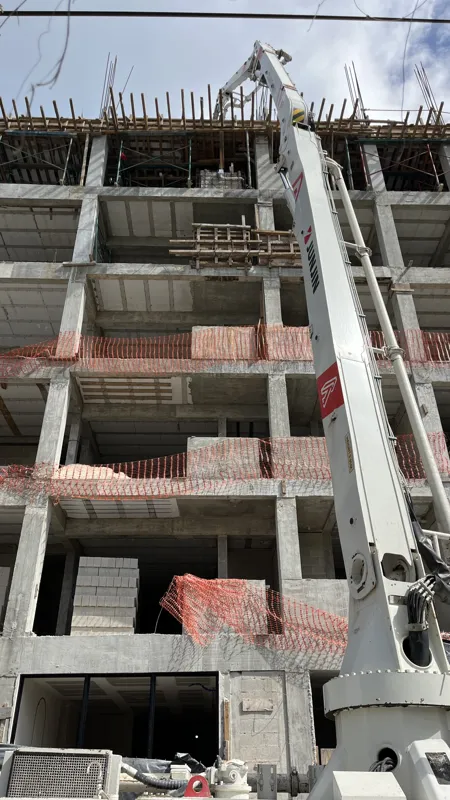 A concrete pump truck and mixer truck actively pouring concrete onto the 6th level of the Maia development building, which features a multi-story concrete structure with rebar visible at the top and orange safety netting on the lower floors, under...