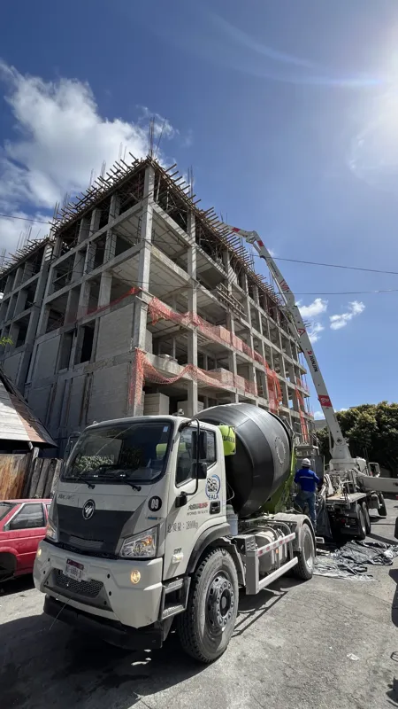 A concrete pump truck and mixer truck actively pouring concrete onto the 6th level of the Maia development building, which features a multi-story concrete structure with rebar visible at the top and orange safety netting on the lower floors, under...
