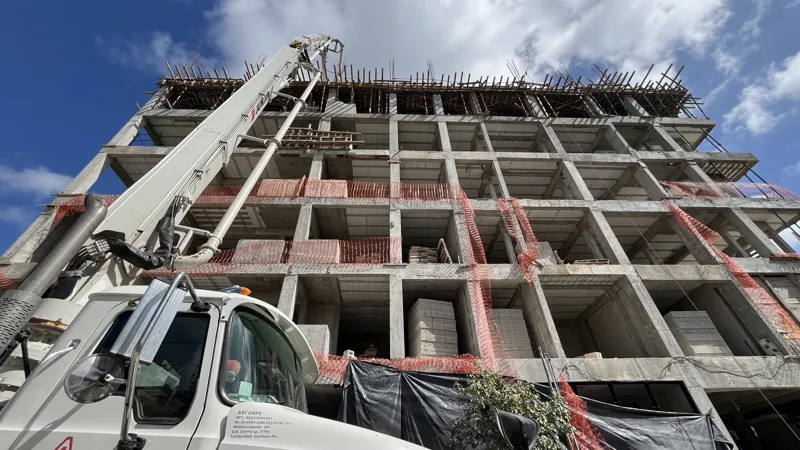 A concrete pump truck and mixer truck actively pouring concrete onto the 6th level of the Maia development building, which features a multi-story concrete structure with rebar visible at the top and orange safety netting on the lower floors, under...