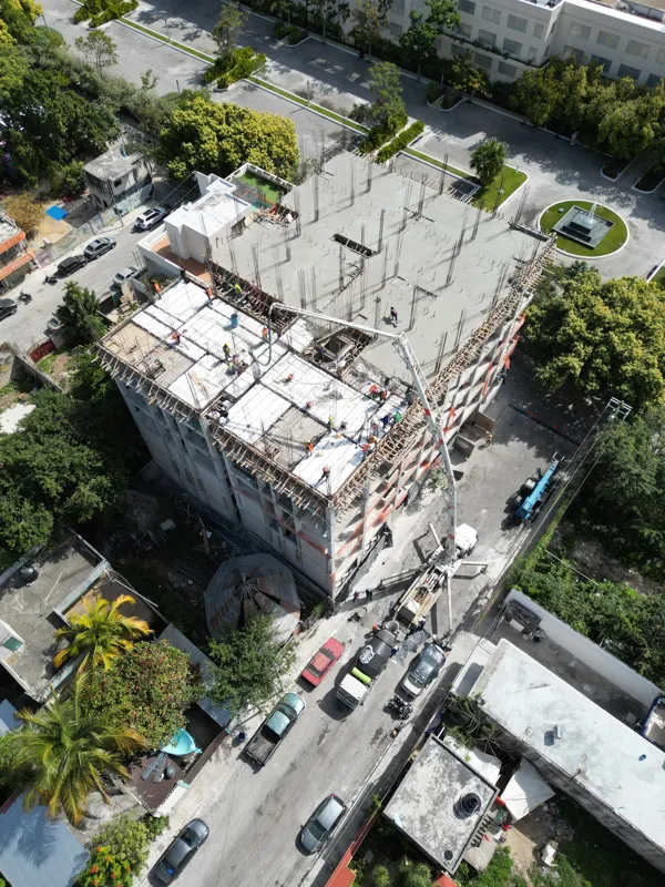 A concrete pump truck and mixer truck actively pouring concrete onto the 6th level of the Maia development building, which features a multi-story concrete structure with rebar visible at the top and orange safety netting on the lower floors, under...