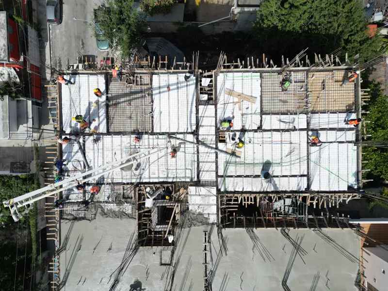 A concrete pump truck and mixer truck actively pouring concrete onto the 6th level of the Maia development building, which features a multi-story concrete structure with rebar visible at the top and orange safety netting on the lower floors, under...