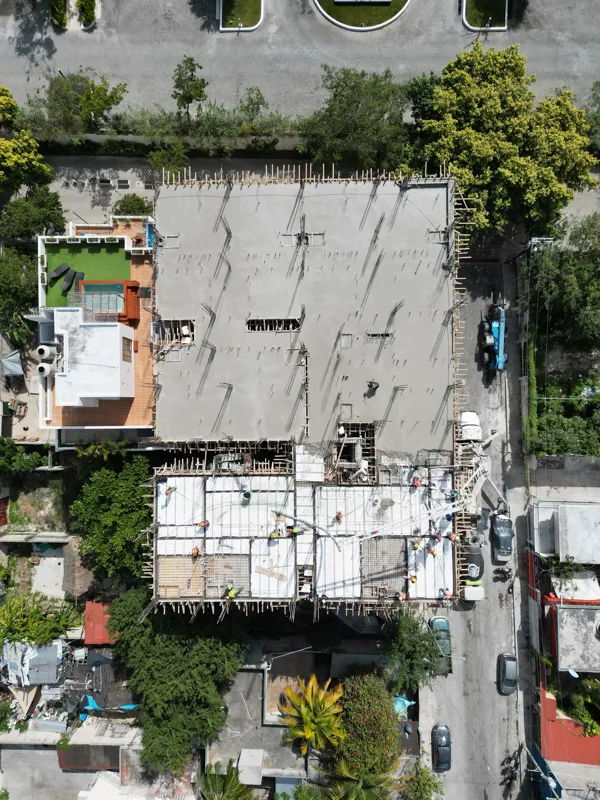 A concrete pump truck and mixer truck actively pouring concrete onto the 6th level of the Maia development building, which features a multi-story concrete structure with rebar visible at the top and orange safety netting on the lower floors, under...
