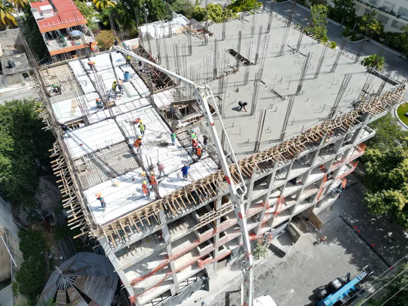 A concrete pump truck and mixer truck actively pouring concrete onto the 6th level of the Maia development building, which features a multi-story concrete structure with rebar visible at the top and orange safety netting on the lower floors, under...