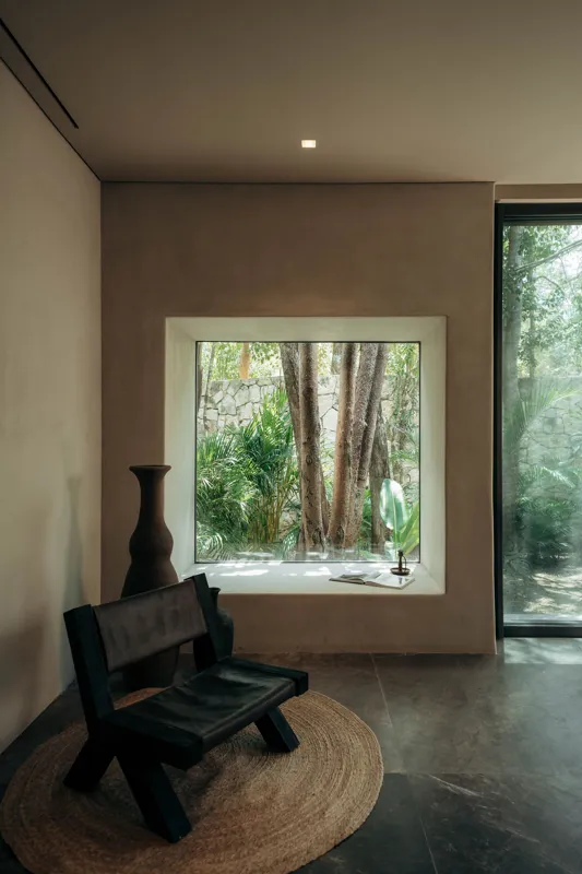 A beautifully designed bedroom in La Reserva Tulum, featuring a king-size bed with neutral-toned bedding, a woven bench at the foot, and two illuminated wall sconces. A large window offers a lush view of a private tropical garden with a stone wall...