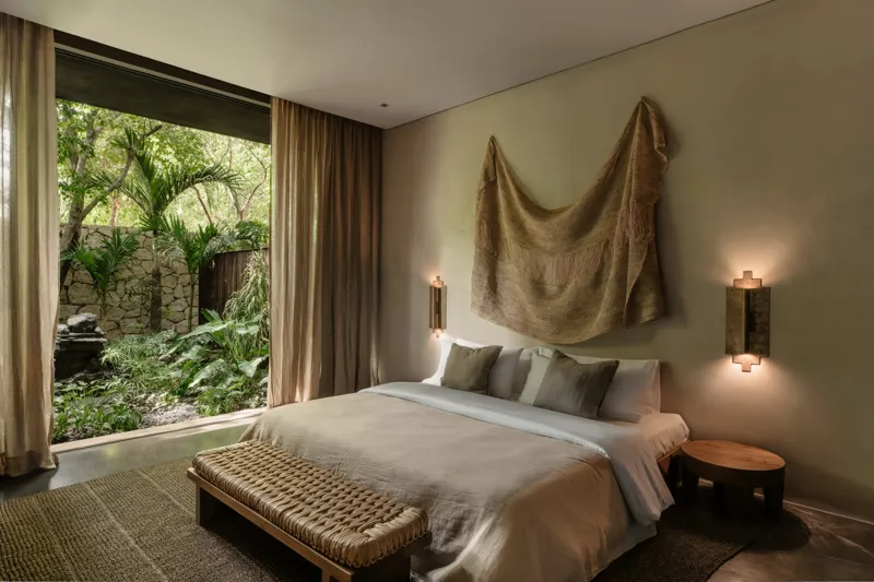 A beautifully designed bedroom in La Reserva Tulum, featuring a king-size bed with neutral-toned bedding, a woven bench at the foot, and two illuminated wall sconces. A large window offers a lush view of a private tropical garden with a stone wall...