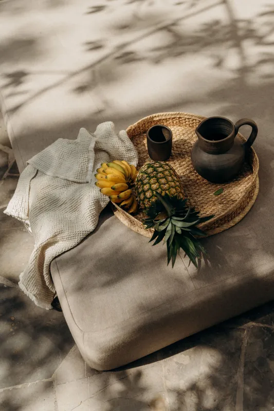 A beautifully designed bedroom in La Reserva Tulum, featuring a king-size bed with neutral-toned bedding, a woven bench at the foot, and two illuminated wall sconces. A large window offers a lush view of a private tropical garden with a stone wall...