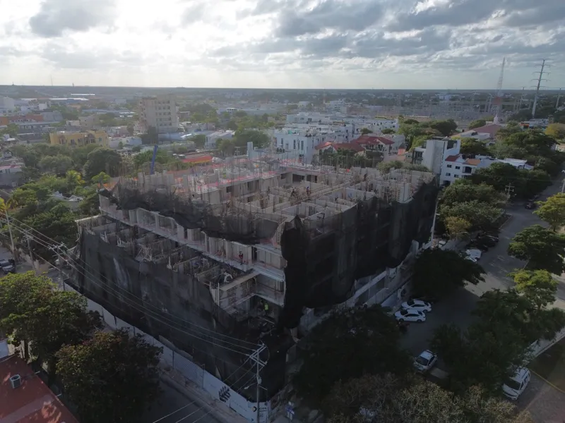 An aerial drone shot captures the ongoing construction of the Costa Celeste apartment complex in Playa del Carmen, showing the multi-story concrete structure with scaffolding and safety netting, surrounded by urban development and lush greenery, w...