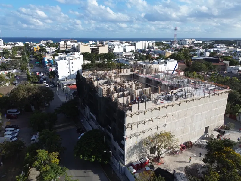 An aerial drone shot captures the ongoing construction of the Costa Celeste apartment complex in Playa del Carmen, showing the multi-story concrete structure with scaffolding and safety netting, surrounded by urban development and lush greenery, w...
