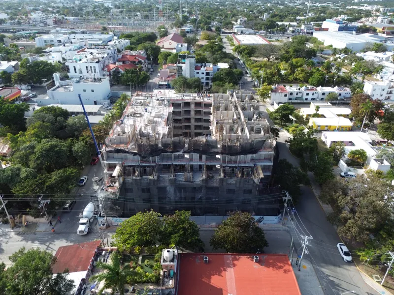 An aerial drone shot captures the ongoing construction of the Costa Celeste apartment complex in Playa del Carmen, showing the multi-story concrete structure with scaffolding and safety netting, surrounded by urban development and lush greenery, w...