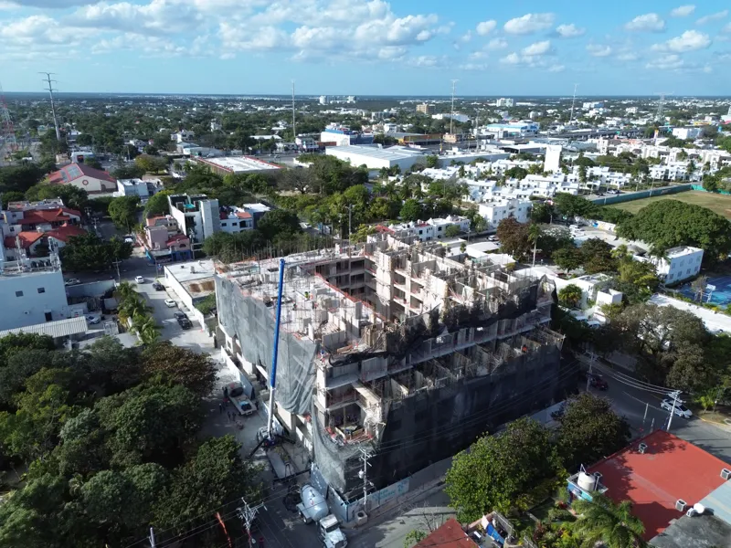 An aerial drone shot captures the ongoing construction of the Costa Celeste apartment complex in Playa del Carmen, showing the multi-story concrete structure with scaffolding and safety netting, surrounded by urban development and lush greenery, w...