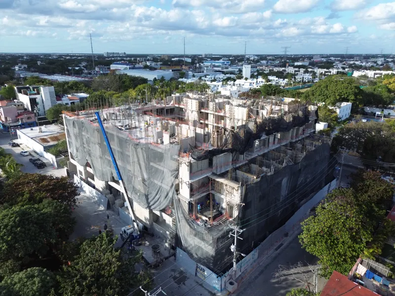 An aerial drone shot captures the ongoing construction of the Costa Celeste apartment complex in Playa del Carmen, showing the multi-story concrete structure with scaffolding and safety netting, surrounded by urban development and lush greenery, w...