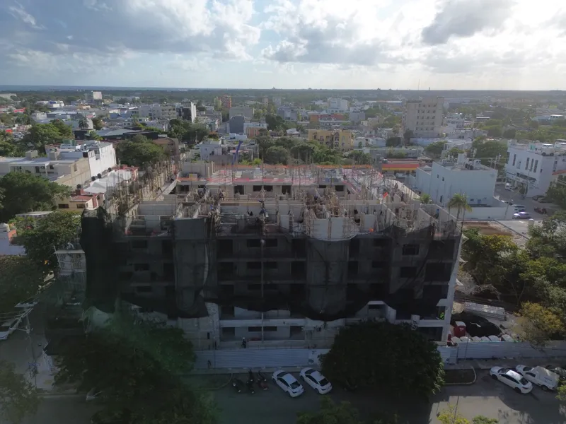 An aerial drone shot captures the ongoing construction of the Costa Celeste apartment complex in Playa del Carmen, showing the multi-story concrete structure with scaffolding and safety netting, surrounded by urban development and lush greenery, w...