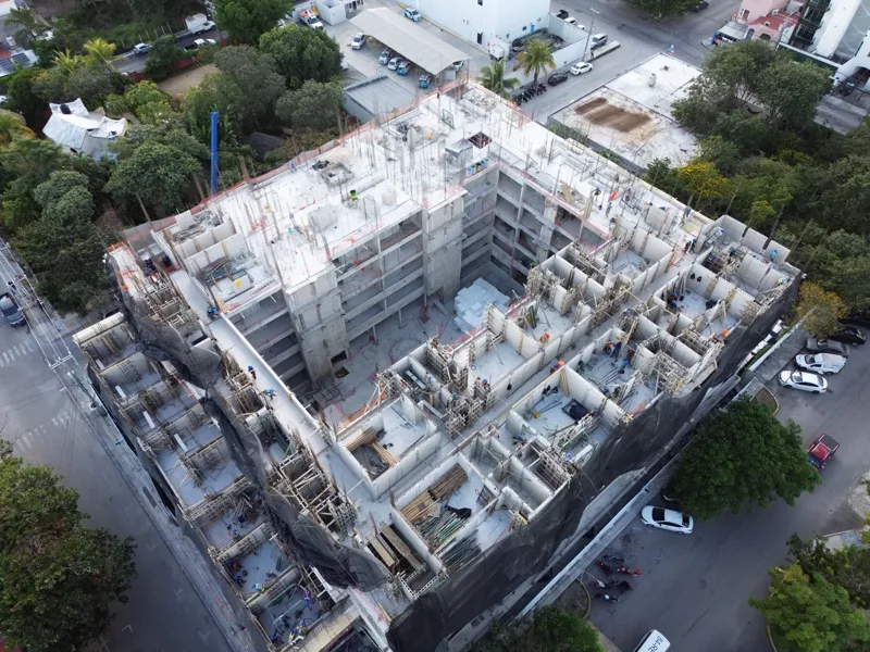 An aerial drone shot captures the ongoing construction of the Costa Celeste apartment complex in Playa del Carmen, showing the multi-story concrete structure with scaffolding and safety netting, surrounded by urban development and lush greenery, w...