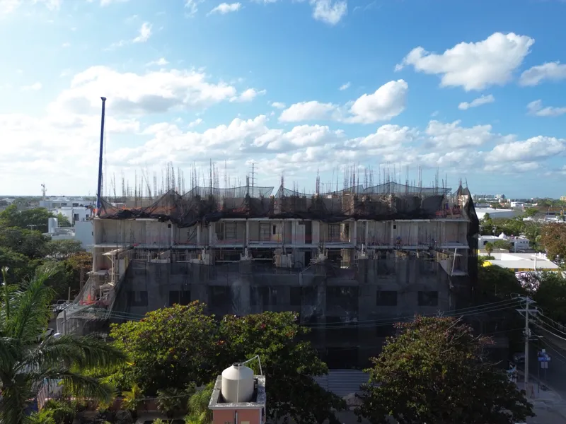 An aerial drone shot captures the ongoing construction of the Costa Celeste apartment complex in Playa del Carmen, showing the multi-story concrete structure with scaffolding and safety netting, surrounded by urban development and lush greenery, w...