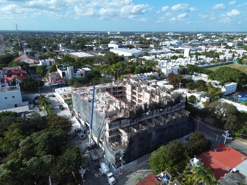 An aerial drone shot captures the ongoing construction of the Costa Celeste apartment complex in Playa del Carmen, showing the multi-story concrete structure with scaffolding and safety netting, surrounded by urban development and lush greenery, w...