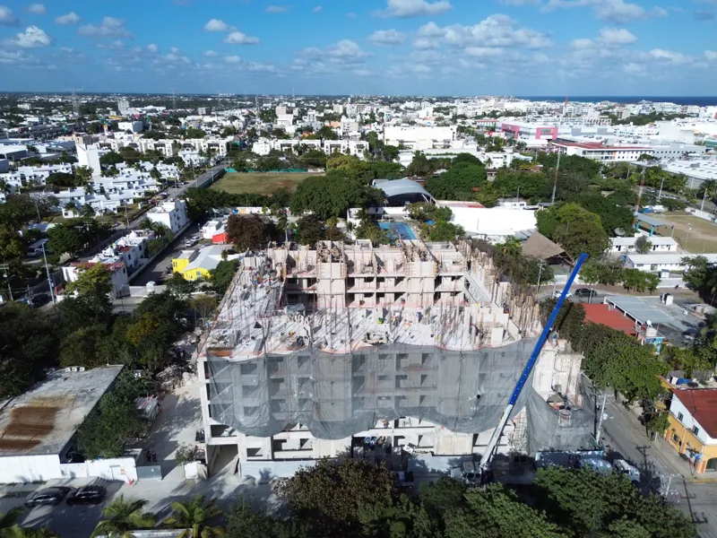 An aerial drone shot captures the ongoing construction of the Costa Celeste apartment complex in Playa del Carmen, showing the multi-story concrete structure with scaffolding and safety netting, surrounded by urban development and lush greenery, w...