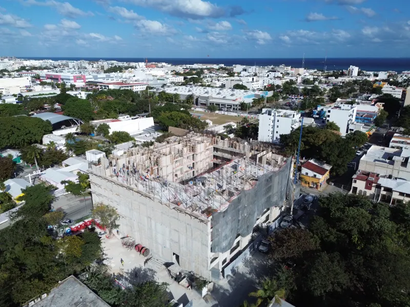 An aerial drone shot captures the ongoing construction of the Costa Celeste apartment complex in Playa del Carmen, showing the multi-story concrete structure with scaffolding and safety netting, surrounded by urban development and lush greenery, w...