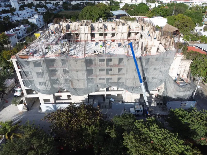 An aerial drone shot captures the ongoing construction of the Costa Celeste apartment complex in Playa del Carmen, showing the multi-story concrete structure with scaffolding and safety netting, surrounded by urban development and lush greenery, w...