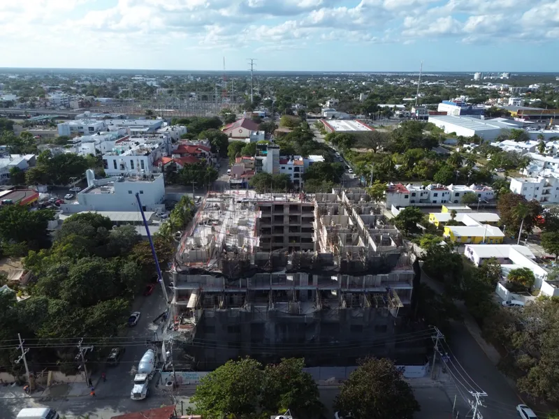 An aerial drone shot captures the ongoing construction of the Costa Celeste apartment complex in Playa del Carmen, showing the multi-story concrete structure with scaffolding and safety netting, surrounded by urban development and lush greenery, w...