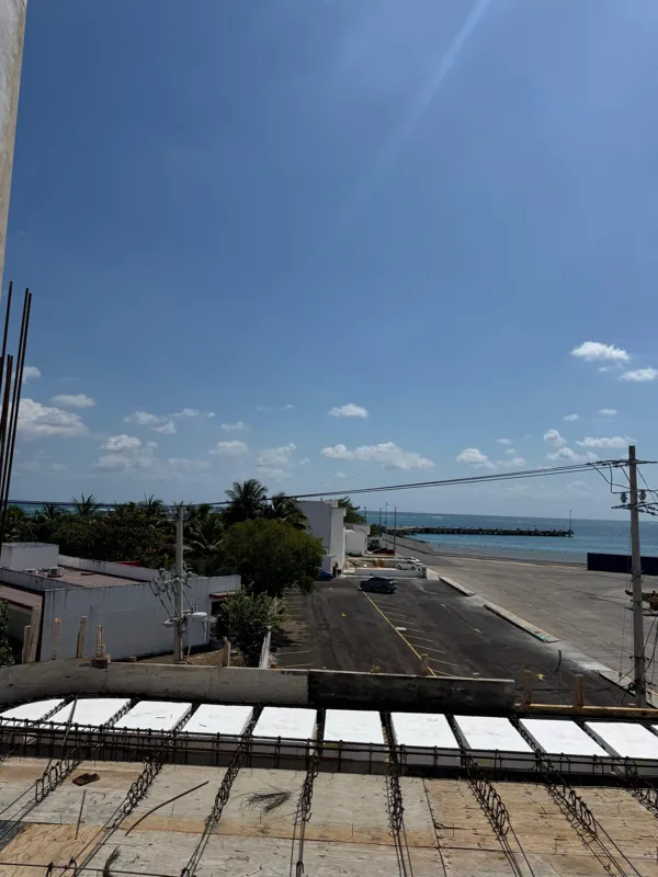 An elevated view from a construction site, showing rebar and formwork for a concrete slab in the foreground. Beyond the construction, a parking lot, green trees, and the clear blue Caribbean Sea with a distant pier are visible under a bright sky w...