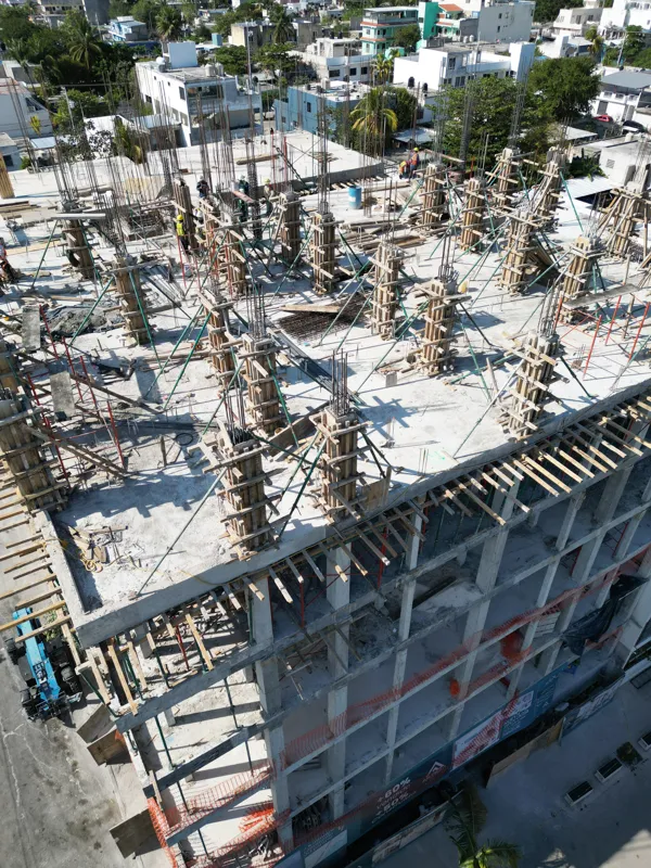 Aerial view of the Maia development construction site in Playa del Carmen, showing multiple concrete columns with rebar and wooden formwork being erected on the upper levels, indicating active progress on the 7th floor, with construction workers v...
