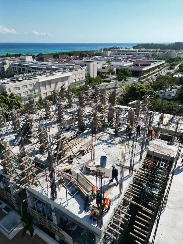 Aerial view of the Maia development construction site in Playa del Carmen, showing multiple concrete columns with rebar and wooden formwork being erected on the upper levels, indicating active progress on the 7th floor, with construction workers v...