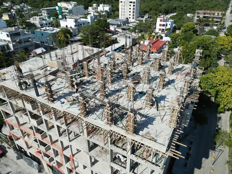 Aerial view of the Maia development construction site in Playa del Carmen, showing multiple concrete columns with rebar and wooden formwork being erected on the upper levels, indicating active progress on the 7th floor, with construction workers v...