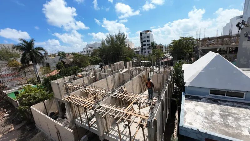 An elevated wide shot of the Piedra de Mar construction site in Puerto Morelos, Mexico, under a bright blue sky. The image shows the multi-section development with concrete block walls forming the ground floor and partially the first floor of seve...
