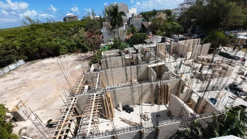 An elevated wide shot of the Piedra de Mar construction site in Puerto Morelos, Mexico, under a bright blue sky. The image shows the multi-section development with concrete block walls forming the ground floor and partially the first floor of seve...