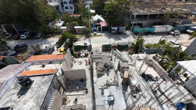 An elevated wide shot of the Piedra de Mar construction site in Puerto Morelos, Mexico, under a bright blue sky. The image shows the multi-section development with concrete block walls forming the ground floor and partially the first floor of seve...