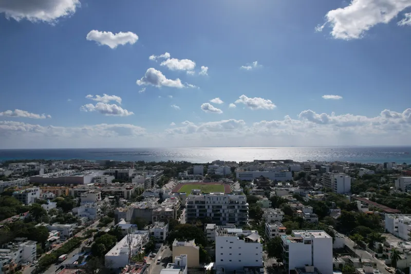 Aerial view of the fully constructed Ceiba at 25 luxury residential building in Playa del Carmen, showcasing its modern white facade, multiple levels of balconies with glass railings, and a prominent rooftop featuring a large, blue-tiled swimming ...