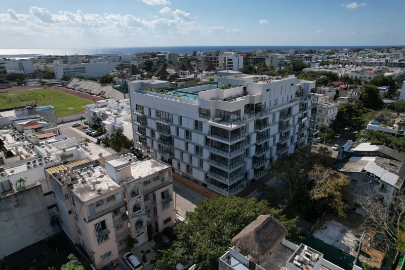 Aerial view of the fully constructed Ceiba at 25 luxury residential building in Playa del Carmen, showcasing its modern white facade, multiple levels of balconies with glass railings, and a prominent rooftop featuring a large, blue-tiled swimming ...