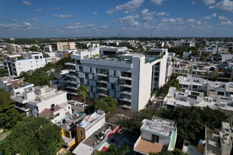 Aerial view of the fully constructed Ceiba at 25 luxury residential building in Playa del Carmen, showcasing its modern white facade, multiple levels of balconies with glass railings, and a prominent rooftop featuring a large, blue-tiled swimming ...