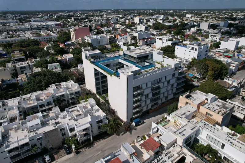 Aerial view of the fully constructed Ceiba at 25 luxury residential building in Playa del Carmen, showcasing its modern white facade, multiple levels of balconies with glass railings, and a prominent rooftop featuring a large, blue-tiled swimming ...