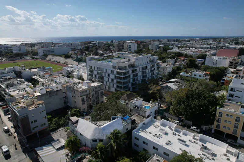 Aerial view of the fully constructed Ceiba at 25 luxury residential building in Playa del Carmen, showcasing its modern white facade, multiple levels of balconies with glass railings, and a prominent rooftop featuring a large, blue-tiled swimming ...