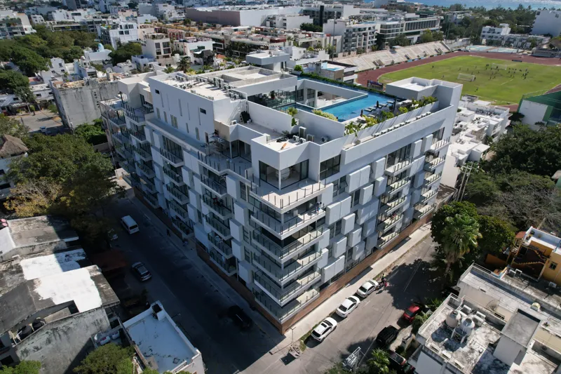 Aerial view of the fully constructed Ceiba at 25 luxury residential building in Playa del Carmen, showcasing its modern white facade, multiple levels of balconies with glass railings, and a prominent rooftop featuring a large, blue-tiled swimming ...