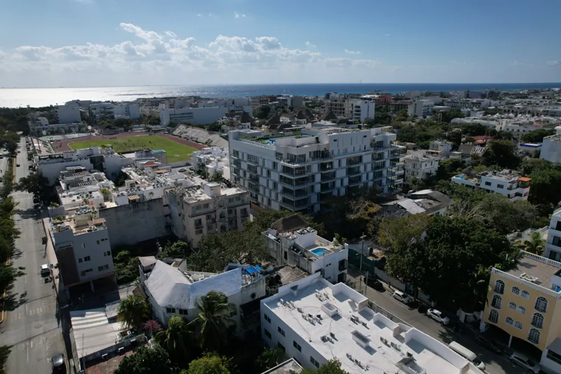 Aerial view of the fully constructed Ceiba at 25 luxury residential building in Playa del Carmen, showcasing its modern white facade, multiple levels of balconies with glass railings, and a prominent rooftop featuring a large, blue-tiled swimming ...