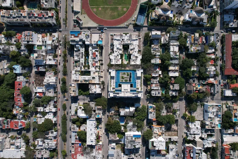 Aerial view of the fully constructed Ceiba at 25 luxury residential building in Playa del Carmen, showcasing its modern white facade, multiple levels of balconies with glass railings, and a prominent rooftop featuring a large, blue-tiled swimming ...